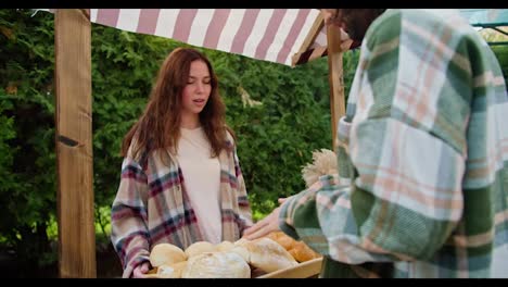 Over-the-shoulder,-a-happy-brunette-guy-in-a-green-checkered-shirt-talks-with-a-girl-who-sells-baked-goods-in-her-shop-during-a-fair-against-the-backdrop-of-green-coniferous-trees-in-summer