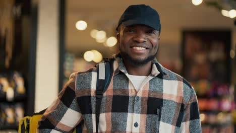 Portrait-of-a-happy-man-with-Black-skin-in-a-checkered-shirt-and-a-black-cap-who-works-as-a-delivery-man-and-carries-a-large-yellow-bag-on-his-shoulders-while-looking-for-the-necessary-products-in-a-modern-supermarket