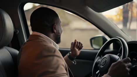 Happy-cheerful-man-Businessman-with-Black-skin-in-a-brown-jacket-drives-a-car-while-sitting-in-a-modern-car-interior-and-dances-during-his-business-trip-in-the-city