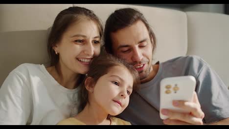 A-happy-brunette-man-in-a-gray-T-shirt-and-his-cheerful-wife,-a-brunette-girl-in-a-white-T-shirt,-together-with-their-daughter,-a-little-girl-in-a-yellow-dress,-take-a-selfie-and-look-at-the-screen-of-a-White-phone-while-sitting-on-a-large-light-brown-sofa