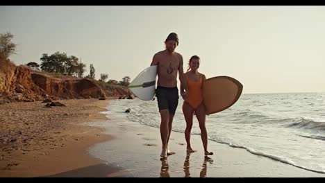 A-blond-man-in-black-shorts-walks-and-chats-with-his-girlfriend-in-an-orange-swimsuit,-they-hold-hands-and-carry-surfboards-while-walking-along-the-sandy-rocky-seashore-in-the-morning-at-Sunrise