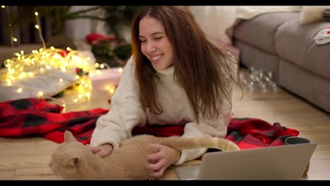 Happy-brunette-girl-in-a-White-sweater-lies-on-the-floor-on-a-Red-blanket-near-her-cream-cat,-strokes-it-and-watches-a-movie-on-a-gray-laptop-in-a-cozy-room-decorated-in-Christmas-style