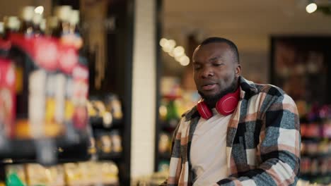 A-happy-man-with-Black-skin-color-in-a-checkered-shirt-and-in-red-wireless-headphones-dances-among-the-counters-in-a-modern-grocery-store-and-has-fun-during-his-shopping