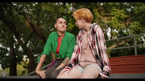 A-LGBT-couple,-a-stole-girl-with-short-hair-in-a-plaid-shirt-and-a-girl-with-short-green-hair-in-a-Green-shirt,-are-sitting-on-a-fence-in-the-park-and-flirting-with-each-other.-Two-lesbian-girls-on-a-date-in-the-park