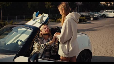 A-blonde-girl-in-a-White-sweatshirt-stands-near-her-blonde-boyfriend-in-a-checkered-shirt-who-sits-in-a-White-convertible-and-kisses-his-girlfriend’s-hand.-Happy-couple-near-their-Convertible-with-surfboards-in-the-city