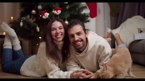 Portrait-of-a-happy-couple,-a-guy-and-a-brunette-girl-in-white-sweaters-lie-on-the-floor-near-their-cream-colored-cat-near-the-Christmas-Tree-in-an-apartment-that-is-decorated-in-the-spirit-of-Christmas