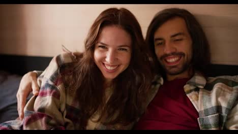 A-happy-brunette-girl-in-a-plaid-shirt-together-with-her-brunette-boyfriend-with-stubble-are-watching-a-movie-on-a-laptop-while-lying-on-a-bed-in-a-trailer-in-a-camp-during-their-picnic-outside-the-city-in-the-summer