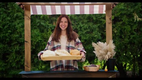 A-happy-brunette-girl-holds-a-tray-with-various-delicious-pastries-in-her-hands-during-her-work-in-a-bread-shop-against-the-backdrop-of-green-coniferous-trees-in-summer