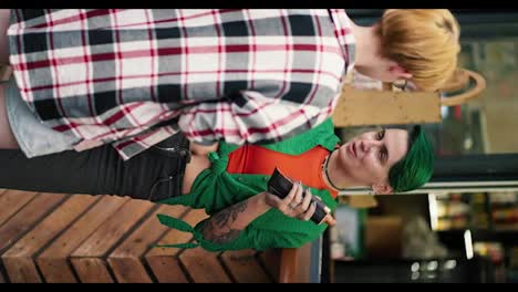 Vertical-video:-blonde-girl-with-a-short-haircut-in-a-turn-off-shirt-communicates-with-her-girlfriend-with-green-short-hair-in-a-green-shirt-who-holds-a-hot-dog-in-her-hand-near-a-street-cafe-during-her-date-in-the-park