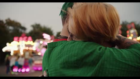 Close-up-shot-of-a-girl-with-short-green-hair-in-a-checkered-green-shirt-and-a-blonde-girl-with-a-short-haircut-in-a-checkered-pink-shirt-hugging-and-looking-at-each-other-during-their-date-in-the-amusement-park