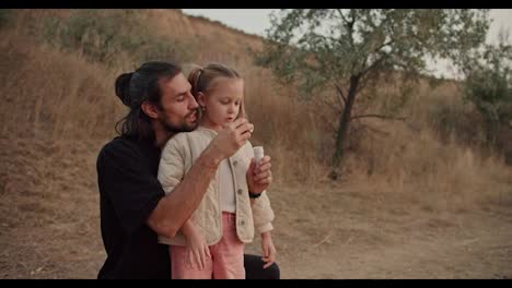 A-little-brunette-girl-blows-soap-bubbles-with-her-father-during-their-picnic-outside-the-city-on-a-summer-evening