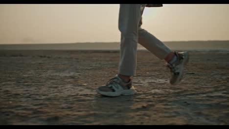 Close-up-shot-of-a-girl-in-gray-shorts-and-sneakers-walking-along-a-deserted-seashore-during-her-vacation-outside-the-city-in-a-summer-evening