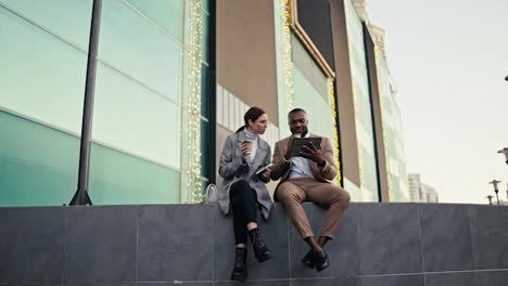 A-businesswoman-girl-wearing-round-glasses-and-a-gray-jacket-sits-on-a-large-gray-building-and-communicates-with-her-colleague-a-man-with-Black-skin-while-they-are-drinking-coffee-and-presenting-their-plans-on-a-black-tablet-in-the-city