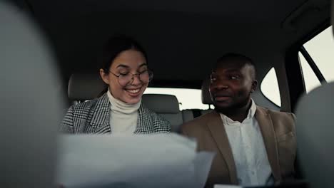 Happy-brunette-girl-in-round-glasses-communicates-with-her-colleague-a-man-with-Black-skin-in-a-brown-jacket-while-they-review-their-plans-and-thoughts-on-paper-while-traveling-in-a-modern-salon-in-a-car-outside-the-city