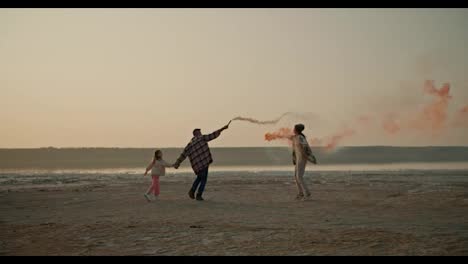 A-happy-middle-aged-man-in-a-checkered-shirt-together-with-his-little-daughter-and-his-wife,-a-brunette-woman-in-a-checkered-green-shirt,-are-holding-fireworks-in-their-hands-that-produce-green-and-red-smoke-during-their-relaxation-and-fun-outside-the-city-on-the-deserted-seashore-in-the-summer-even