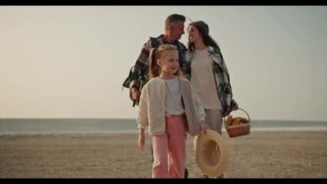 A-little-blonde-girl-in-a-white-jacket-and-pink-pants-walks-along-a-deserted-path-and-behind-her-are-her-parents,-a-brunette-man-and-a-brunette-girl-in-a-Green-checkered-shirt-who-carries-a-basket-of-food-during-their-picnic-outside-the-city