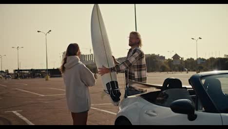 A-blond-guy-in-a-plaid-shirt-and-glasses-gives-his-blonde-girlfriend-in-a-white-sweatshirt-a-white-surfboard-from-his-white-Convertible-and-takes-his-one-on-the-Big-Square-in-the-city