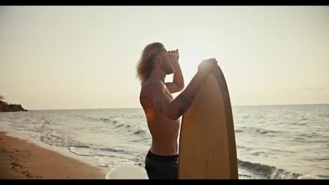 A-blond-man-with-a-beard-with-a-bare-torso-leans-with-one-hand-on-his-surfboard-and-with-the-other-he-covers-his-eyes-from-the-sun-and-looks-into-the-sea-while-standing-on-the-shore-in-the-morning-at-Sunrise