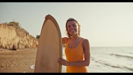 Portrait-of-a-happy-young-blonde-girl-in-an-orange-swimsuit-who-stands-and-looks-at-the-camera-holding-a-wooden-yellow-surfboard-on-a-sandy-rocky-beach-near-the-sea-in-the-morning