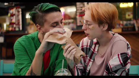 Two-lesbian-girls-with-short-haircuts-in-bright-plaid-shirts-feed-each-other-hot-dogs-in-a-street-cafe-in-the-park-during-their-date.-Two-lesbian-girls-during-their-LGBT-date-in-the-park-in-the-summer