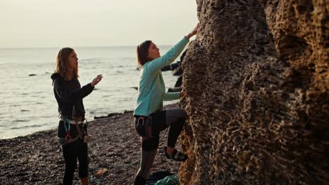 A-blonde-girl-with-a-bob-hairstyle-in-a-blue-sweater-and-black-sweatpants-climbs-onto-a-rock-in-special-equipment,-and-a-blonde-girl-in-black-clothes-insures-her-climbing-student,-stands-below-and-guides-her