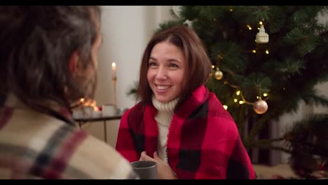 Over-the-shoulder,-a-brunette-guy-in-a-checkered-shirt-is-talking-to-his-brunette-girlfriend-in-a-white-sweater-and-a-red-checkered-shirt-who-is-drinking-a-hot-drink-from-a-gray-mug-near-a-green-New-Year-tree-in-a-cozy-room-in-winter