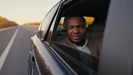 A-suspicious-Black-man-with-a-dissatisfied-face-in-a-brown-suit-opens-the-rear-window-of-the-car-and-looks-at-the-roadway-during-his-trip-outside-the-city.-Dissatisfied-pensive-male-businessman-looking-out-the-window