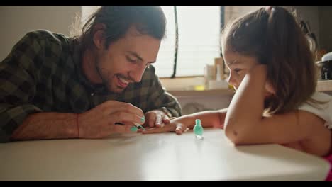 Un-Hombre-Moreno-Con-Una-Camisa-A-Cuadros-Verde-Le-Hace-La-Manicura-Con-Esmalte-Verde-A-Su-Pequeña-Hija,-Una-Niña-Morena-Con-Un-Vestido-Rosa,-En-La-Cocina-Sobre-La-Mesa.