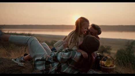 A-little-blonde-girl-in-a-white-jacket-runs-to-her-parents,-a-brunette-man-and-a-girl-in-a-green-checkered-shirt-jumps-on-them-and-knocks-them-onto-the-mat-during-a-picnic-outside-the-city-on-a-summer-evening