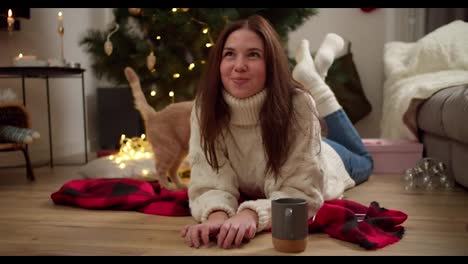 Happy-and-playful-brunette-girl-in-a-White-sweater-plays-with-her-cream-colored-cat-lying-on-a-Red-blanket-in-a-cozy-room-decorated-in-the-New-Year-style