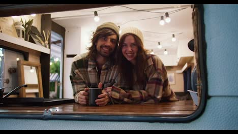 Portrait-of-a-happy-brunette-girl-in-a-white-hat-in-a-plaid-shirt-together-with-her-brunette-boyfriend-in-a-plaid-shirt-drinking-tea-in-a-trailer-and-relaxing-during-their-picnic-in-a-camp-outside-the-city-in-the-summer