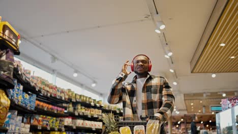 A-happy-man-with-Black-skin-in-a-plaid-shirt-with-a-beard-and-in-red-wireless-headphones-dances-and-rolls-a-cart-among-the-counters-in-a-modern-grocery-store