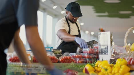 A-Black-man-in-a-white-T-shirt-and-a-black-apron-in-a-black-cap-sorts-vegetables-on-the-counter-in-a-modern-grocery-store.-Employees-of-a-large-modern-store-lay-out-goods-on-the-counter-during-their-duty