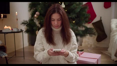 A-brunette-girl-in-white-light-looks-and-does-something-on-her-white-phone-while-sitting-near-a-large-pink-gift-and-a-New-Year-tree-at-home-in-a-cozy-apartment-decorated-in-the-style-of-the-New-Year