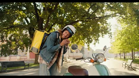 Courier-guy-with-curly-hair-in-a-denim-shirt-puts-on-a-white-motorcycle-helmet-and-looks-out-the-side-window-of-his-moped-along-with-a-big-yellow-one-in-a-sunny-city-park-in-summer