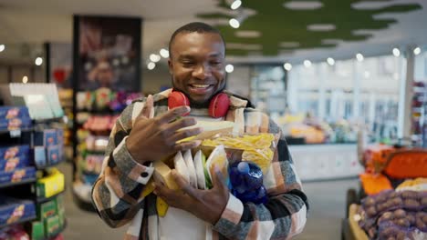 A-happy-and-smiling-man-with-Black-skin-in-a-checkered-shirt-and-red-wireless-headphones-carries-a-bunch-of-groceries-on-his-chest-holding-with-both-hands-during-his-walk-and-shopping-in-a-modern-supermarket