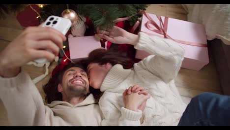 A-brunette-guy-and-his-girlfriend-in-a-white-sweater-lie-on-the-floor-and-take-a-selfie-near-the-New-Year-tree-and-pink-gifts-in-a-cozy-room-in-winter