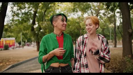 Front-view-of-two-lesbian-girls-with-bright-short-hair-in-shirts-walk-through-the-park-during-their-date-and-communicate