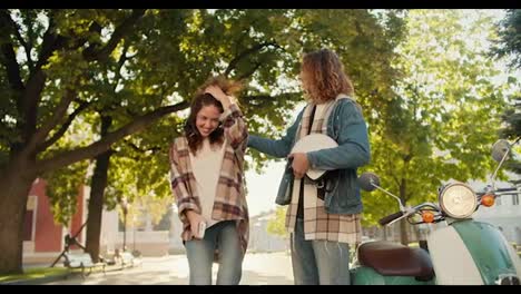 Happy-couple,-a-guy-with-long-curly-hair-in-a-denim-shirt-and-his-brunette-girlfriend-are-dancing-and-hugging-near-their-green-moped-in-a-summer-city-park