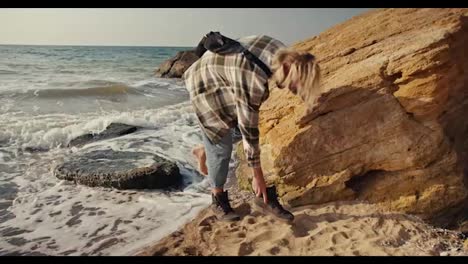 Ein-Blonder-Typ-In-Einem-Karierten-Hemd-Springt-Von-Einem-Stein-Zum-Strand-Und-Versucht,-Seine-Schuhe-Am-Felsigen-Ufer-Des-Strandes-In-Der-Nähe-Des-Meeres-Am-Morgen-Anzuziehen