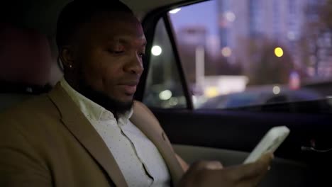 A-confident-man-with-Black-skin-and-a-short-haircut-with-a-beard-in-a-brown-suit,-a-businessman,-drives-along-the-evening-city-and-works-on-his-white-smartphone-in-a-modern-car-interior