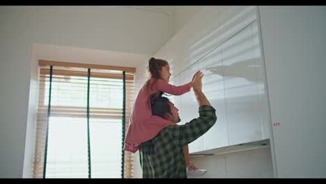 A-little-brunette-girl-in-a-pink-dress-helps-her-brunette-dad-in-a-plaid-shirt-get-an-item-from-the-top-shelves-in-the-kitchen.-A-little-girl-sits-on-her-father's-shoulders-and-takes-an-object-from-a-high-shelf-in-the-kitchen