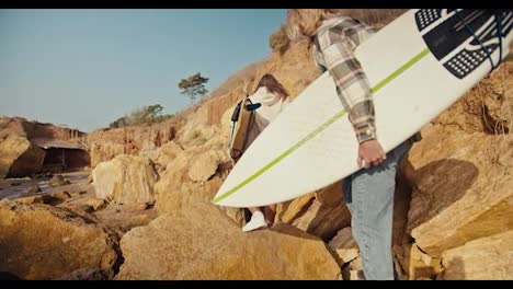 A-blond-guy-in-a-plaid-shirt-and-his-blond-girlfriend-in-a-white-sweatshirt-walk-along-a-rocky-beach-near-the-sea-and-carry-their-surfboards-with-them-on-a-sunny-morning