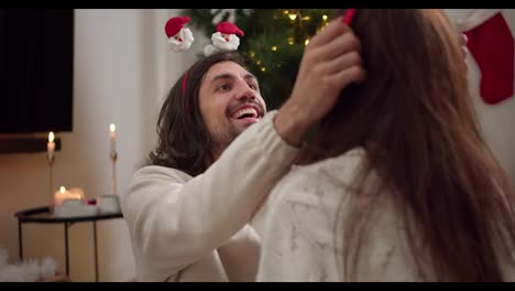 A-happy-brunette-guy-in-a-white-sweater-puts-a-New-Year's-hoop-on-the-head-of-his-brunette-girlfriend-in-a-white-sweater-near-a-green-Christmas-tree-decorated-with-a-yellow-garland-in-a-cozy-winter-home