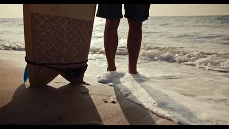 Close-up-shot-of-a-guy-in-shorts-standing-on-the-seashore-and-holding-a-surfboard-near-him-and-small-waves-passing-through-his-legs-and-leaving-sea-foam-on-them-in-the-morning-at-Sunrise