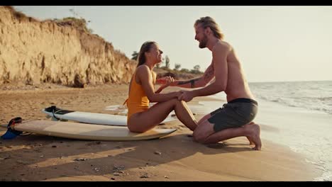 Happy-cheerful-blonde-girl-in-an-orange-swimsuit-holds-hands-with-her-blonde-boyfriend-and-sits-on-a-surfboard-on-the-sandy-seashore-with-rocks-in-the-morning-at-Sunrise