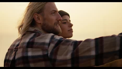 A-blond-guy-with-long-hair-and-a-beard-in-a-checkered-shirt-shows-his-hand-in-the-direction-of-his-girlfriend-and-they-laugh-and-look-at-each-other-in-the-morning-at-Sunrise-near-the-sea