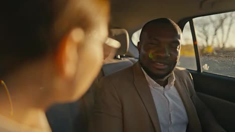 Over-the-shoulder-a-man-with-a-beard-and-Black-skin-in-a-brown-jacket-communicates-with-his-colleague,-a-brunette-girl-with-round-glasses-during-his-trip-to-a-modern-salon-in-a-car-outside-the-city