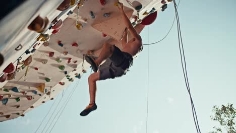 Bottom-view:-an-athletic-guy-in-black-shorts-with-a-sculpted-torso-climbs-up-the-climbing-wall-and-hangs-on-the-ledge-of-the-training-route-of-the-climbing-wall-on-a-sunny-summer-day