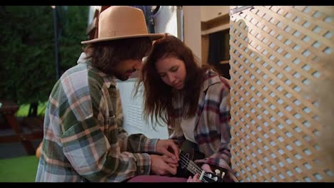 A-brunette-guy-in-a-yellow-hat-helps-his-brunette-girlfriend-in-a-pink-checkered-shirt-play-a-black-ukulele-while-sitting-near-his-trailer-in-a-camp-during-a-picnic-outside-the-city-in-the-summer